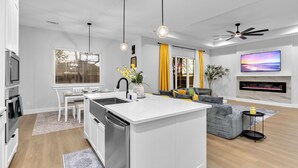 A modern kitchen with a central island, a dining area with a white table and chairs, a living room with a sofa and a TV mounted on the wall. - Luxe 4BR/3BA, 3 King Beds—Near AT&T, DFW, Dallas (Grand Prairie)
