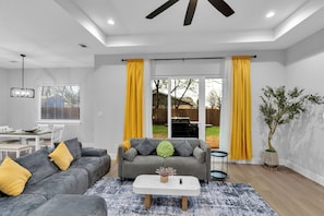 A modern living room with a grey sofa, yellow cushions, a white coffee table, and a large window with yellow curtains. - Luxe 4BR/3BA, 3 King Beds—Near AT&T, DFW, Dallas (Grand Prairie)