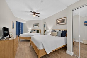 Ein Hotelzimmer mit zwei Betten, einem Schreibtisch mit Computer, einem Deckenventilator und Kunstwerken an den Wänden.