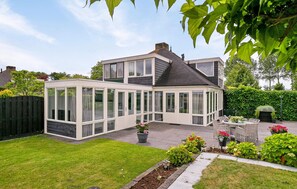 Ett modernt hus i två våningar med stora glasfönster, en uteplats med utemöbler och en välskött trädgård.