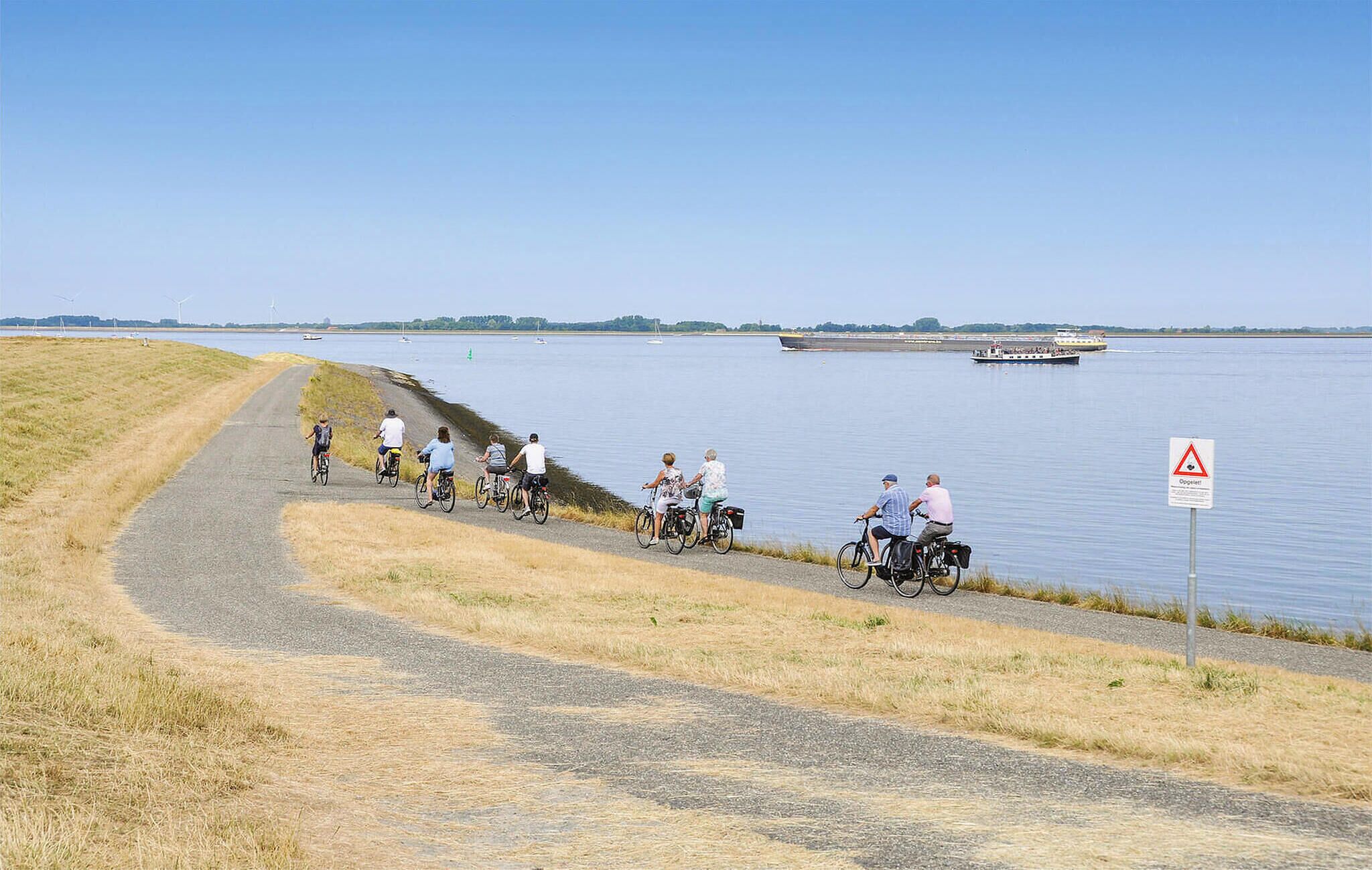 A group of cyclists on a paved path beside a calm body of water, with a warning sign and a boat in the distance.