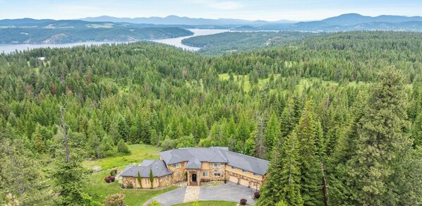 A large house surrounded by dense forest, with a view of a lake and mountains in the distance.