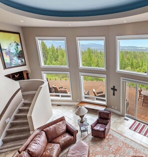 A spacious living area with a staircase, leather furniture, large windows, and a view of a deck. - NW Tuscan Retreat — 180° Vistas & Private Hot Tub (Harrison)