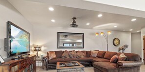 A living room with a sectional sofa, a flat-screen TV, a coffee table, a lamp, a ceiling fan, and a wall clock. - NW Tuscan Retreat — 180° Vistas & Private Hot Tub (Harrison)