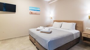Et hotelværelse med en seng, et vægmonteret fladskærms-tv, et maleri af en strandscene og et natbord med en lampe.