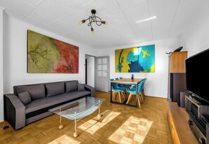 Ein modernes Wohnzimmer mit grauem Sofa, einem Glastisch, einem Holzespeisesaal mit blauen Stühlen und einem großen, abstrakten Gemälde an der Wand.
