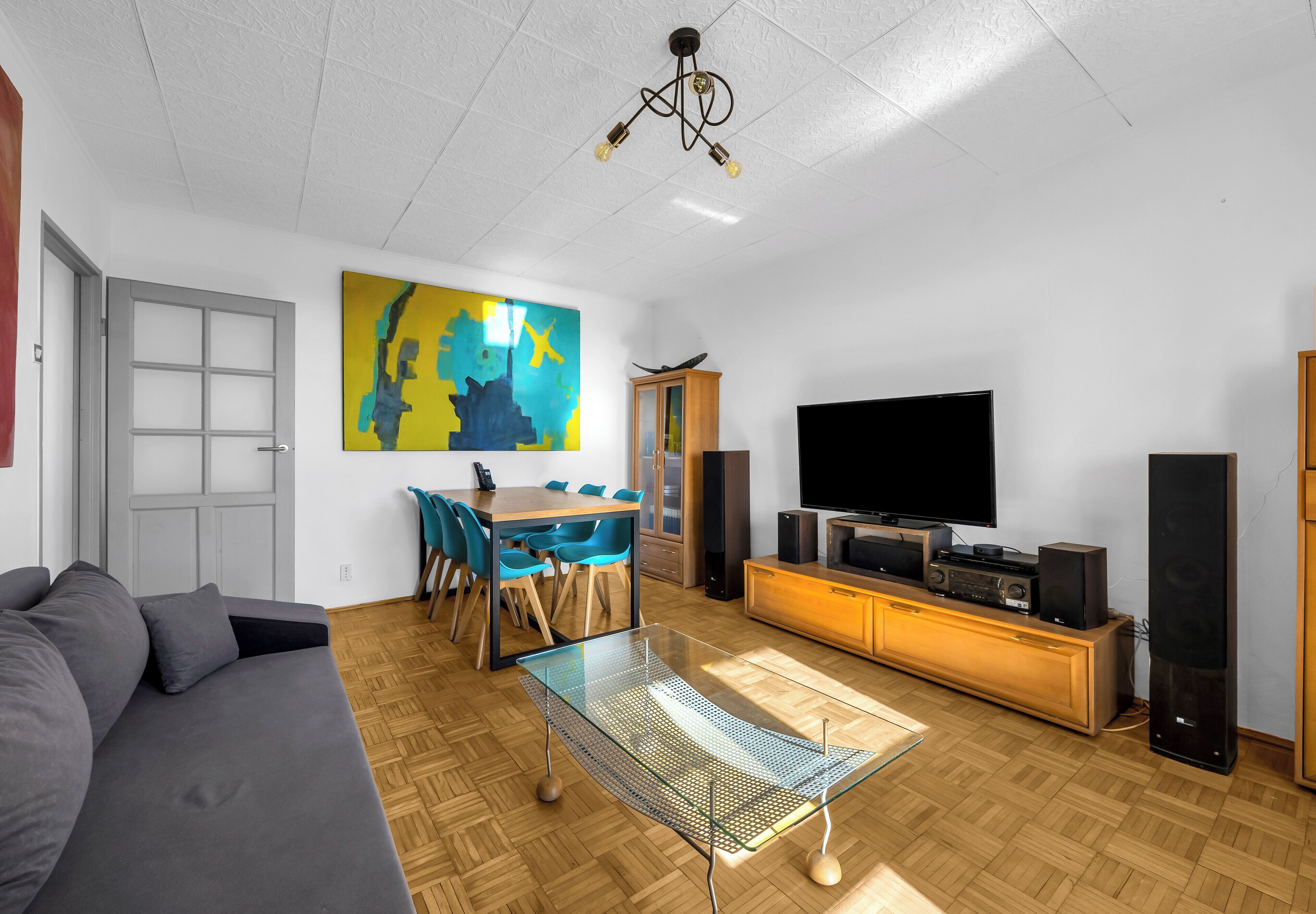 A modern living room with a sofa, a glass coffee table, a dining area with chairs, a TV, and speakers.