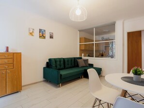 A modern living room with a green sofa, white chairs, a wooden cabinet, a dining table, and a window with a view of a kitchen. - Toruń Urban Comfort Apartment (Toruń)