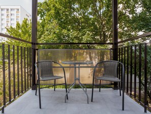 Un balcó amb dues cadires de vimetla i una taula rodona petita, envoltat de baranes metàl·liques i vistes a arbres i edificis.