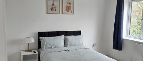 A bedroom with a bed, a nightstand, a lamp, two framed pictures on the wall, and a window with curtains.