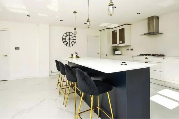 A modern kitchen with a central island, bar stools, pendant lighting, and a clock on the wall.