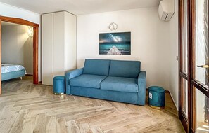 A room with a blue sofa, a painting of a pier, a wardrobe, and a trash bin.
