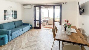 A modern living room with a blue sofa, a wooden dining table with chairs, and a sliding glass door leading to an outdoor area.