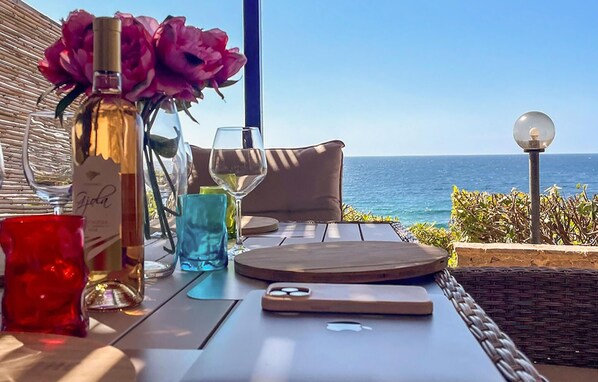 A table with a bottle of perfume, wine glasses, and a smartphone on a balcony overlooking the sea.