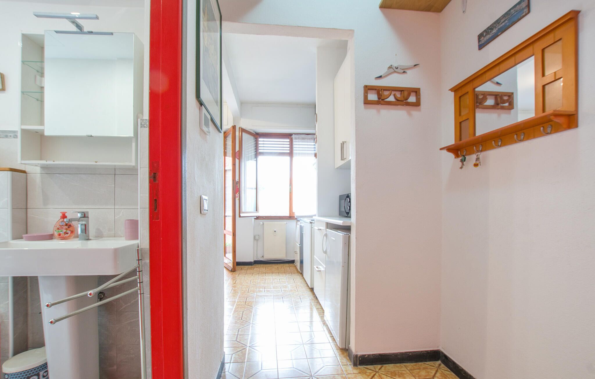 A narrow hallway with a tiled floor leading to a kitchen area. There is a red door on the left, a white sink with a soap dispenser, and a mirror above the sink. The kitchen has white cabinets and a microwave on the countertop.