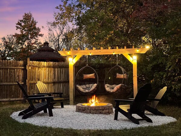A cozy outdoor seating area with two Adirondack chairs, a hanging swing chair, and a fire pit.