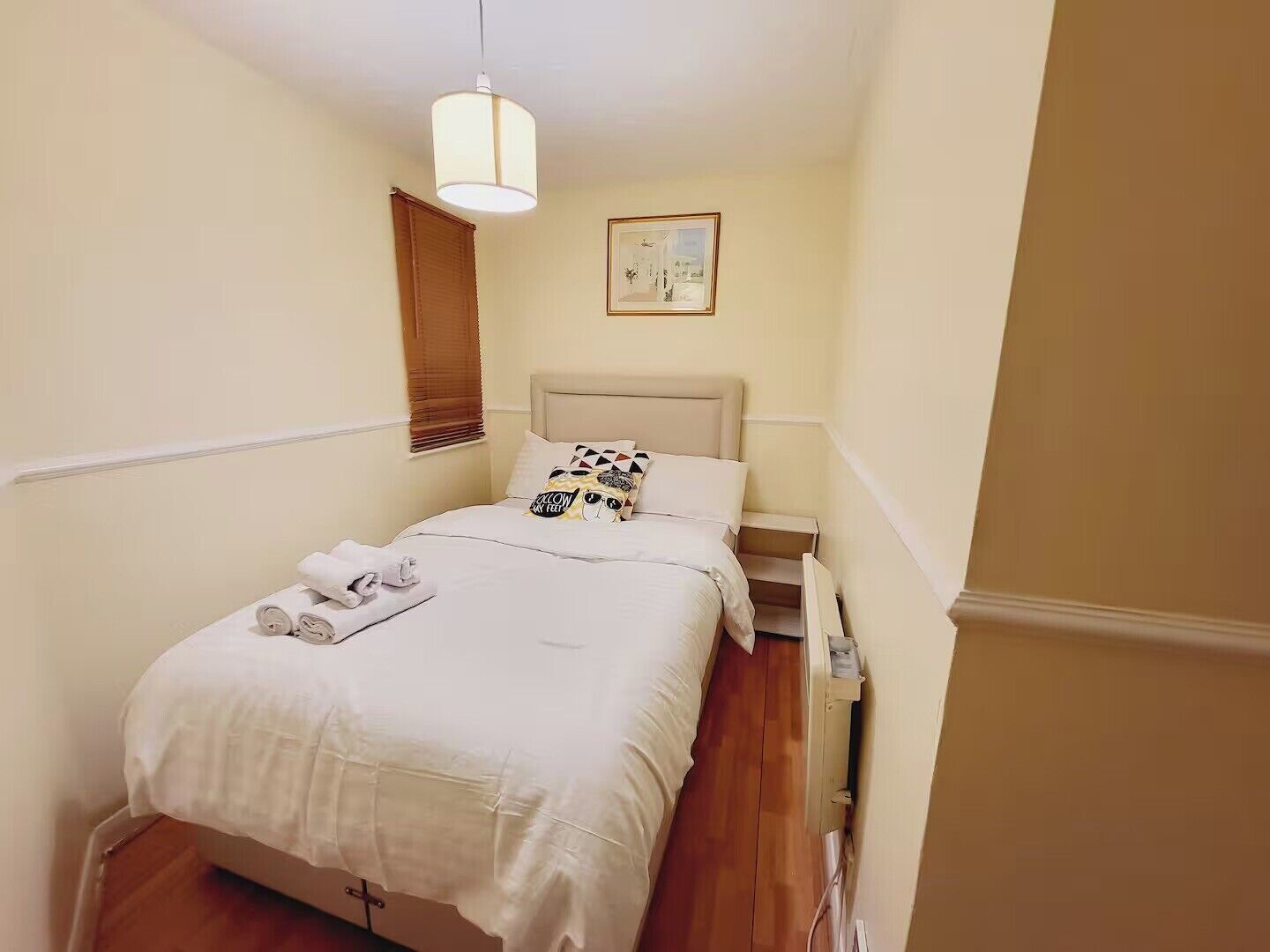 A neatly made bed with white linens, a headboard, and a pillow with a graphic design. There is a small bedside table with a lamp and a framed picture on the wall above the bed.