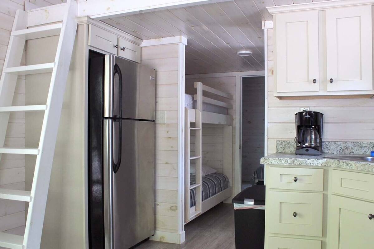 A compact kitchen with a built-in refrigerator, a coffee maker, and a bunk bed in the adjacent room.