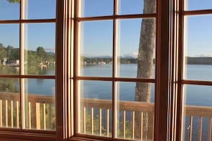 Janela com vista para um lago, uma árvore e montanhas ao longe.