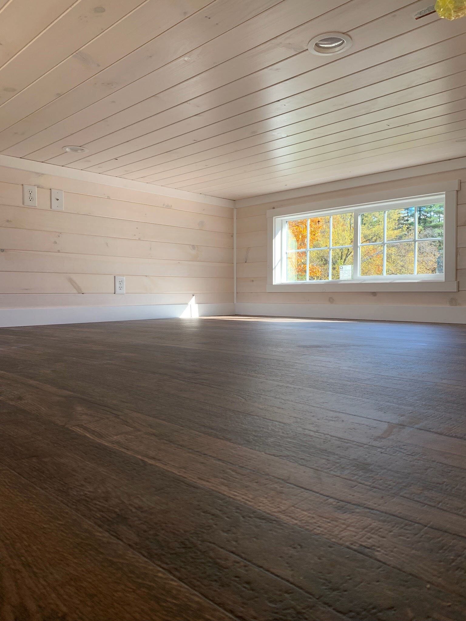 A large, empty room with wooden flooring, a sloped ceiling, and a large window with autumn foliage view.