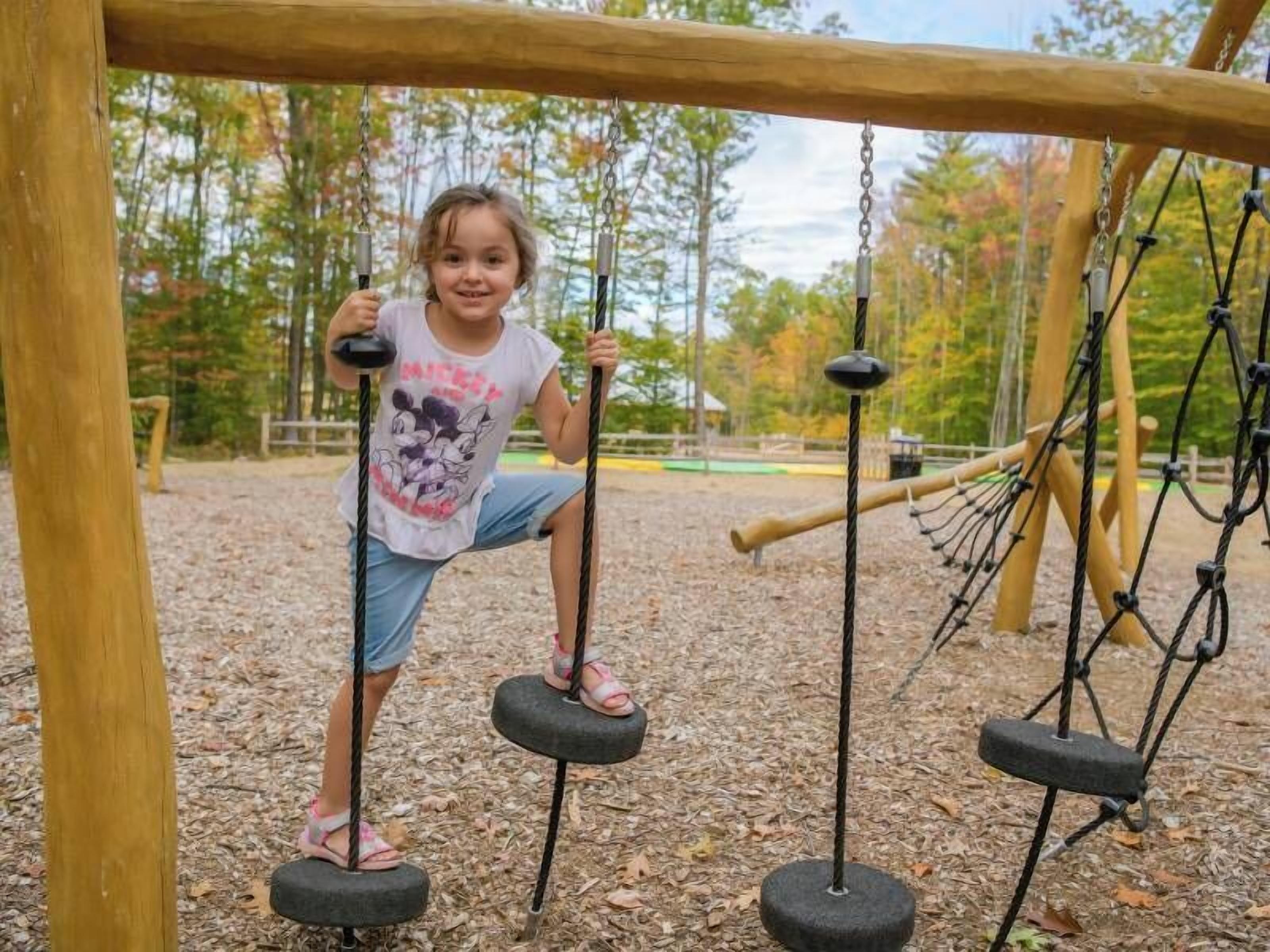 A child on a tire swing at a playground with a wooden structure and trees in the background.