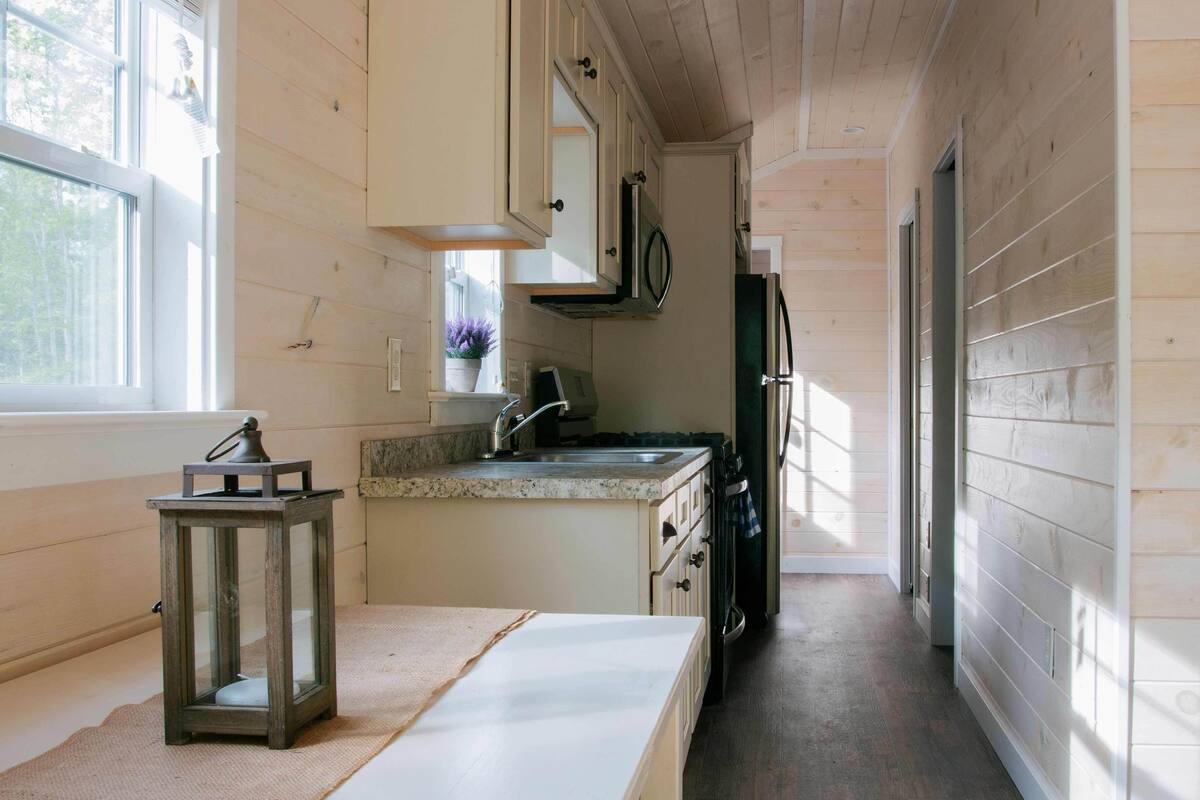 A narrow kitchen with wooden walls, a window, a sink, cabinets, and a refrigerator.