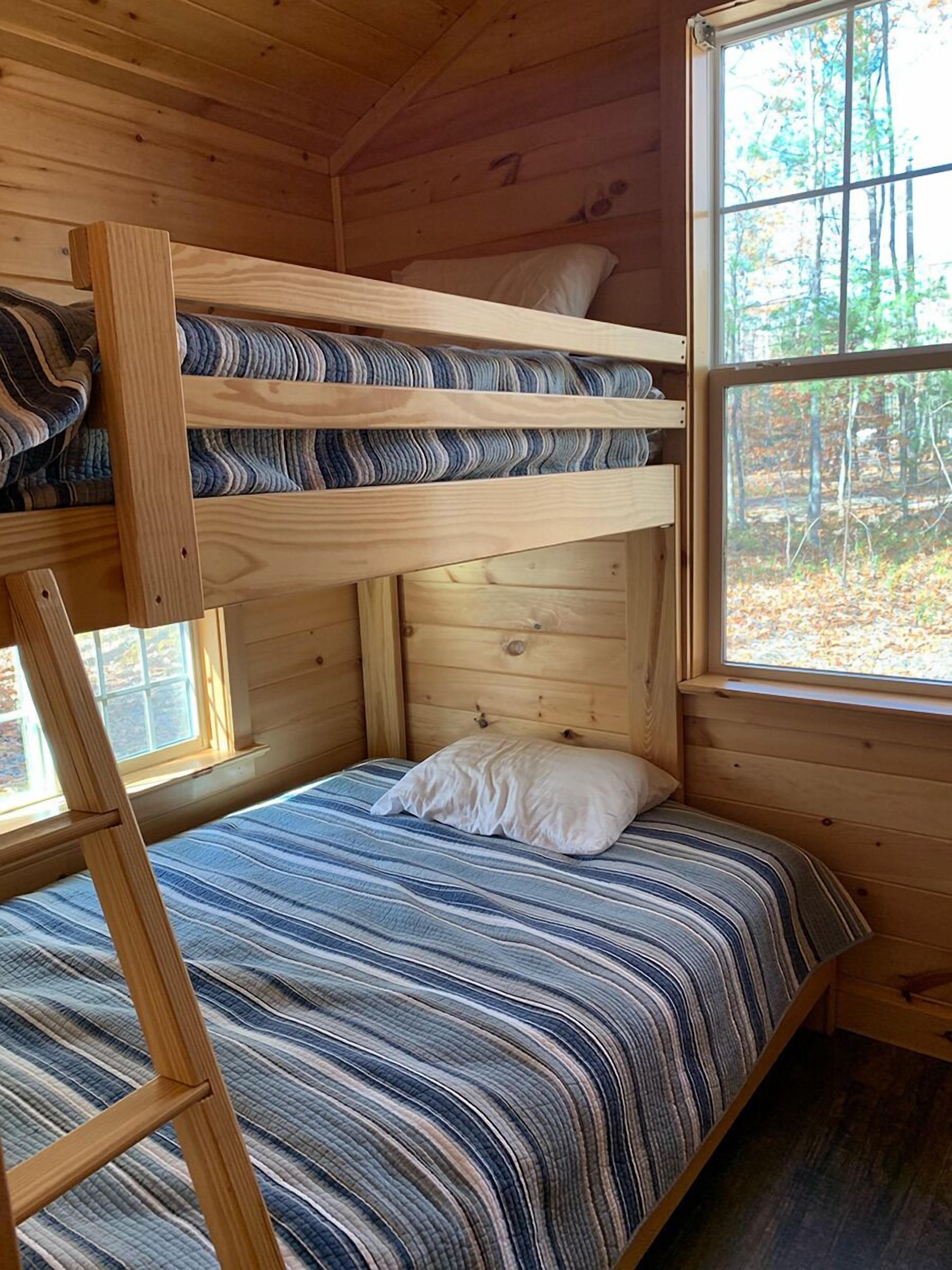 A bunk bed with a striped bedspread, a window with a forest view, and wooden walls.