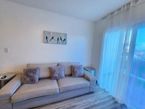 A living room with a beige sofa, a small side table, a painting on the wall, and a large window with sheer curtains. - Modern 2BR/2BA condo, upper floor, ocean breeze, short walk to Playa Chiquita (Sosúa)