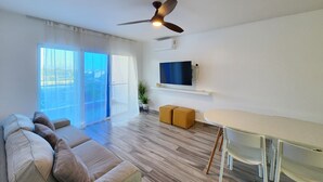 A modern living room with a sofa, a dining table, chairs, a ceiling fan, a TV, and a large window with curtains. - Modern 2BR/2BA condo, upper floor, ocean breeze, short walk to Playa Chiquita (Sosúa)