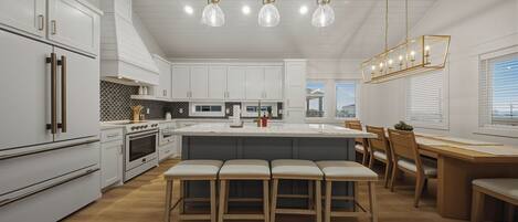 A modern kitchen with white cabinetry, a central island with stools, a dining area with wooden chairs, and pendant lighting.