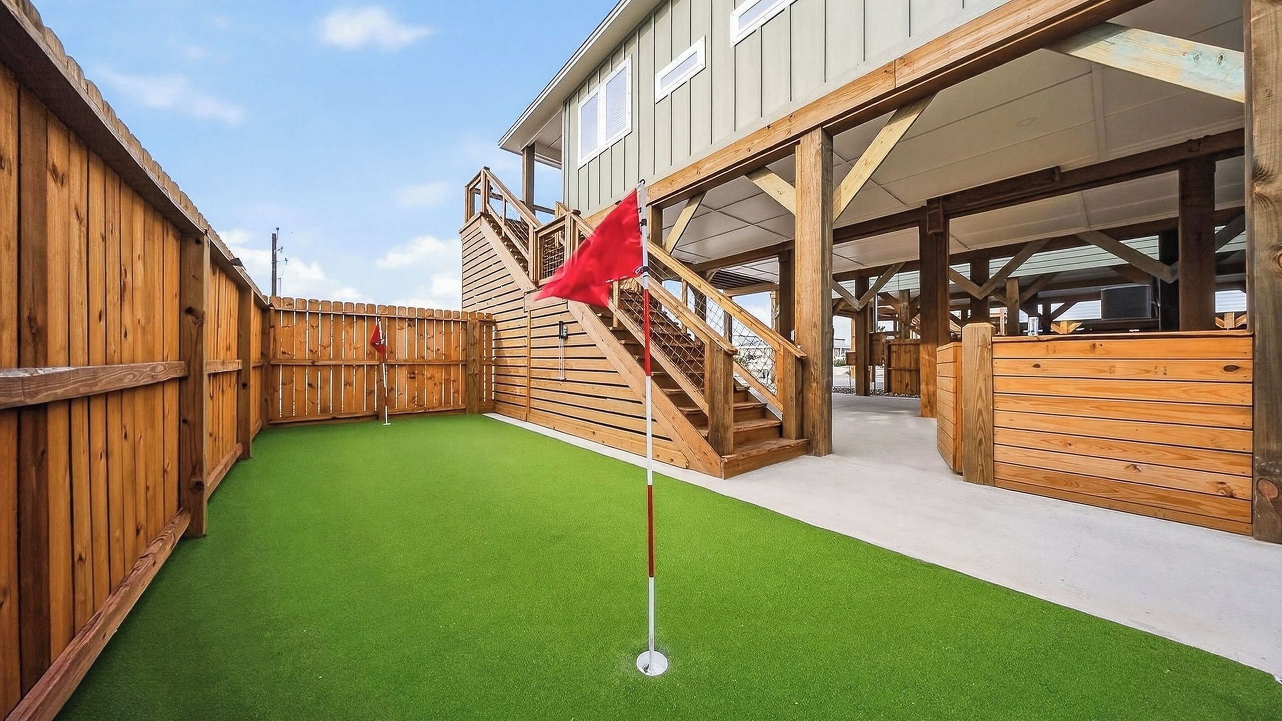 A backyard with artificial green turf, a wooden fence, and a staircase leading to a covered area.
