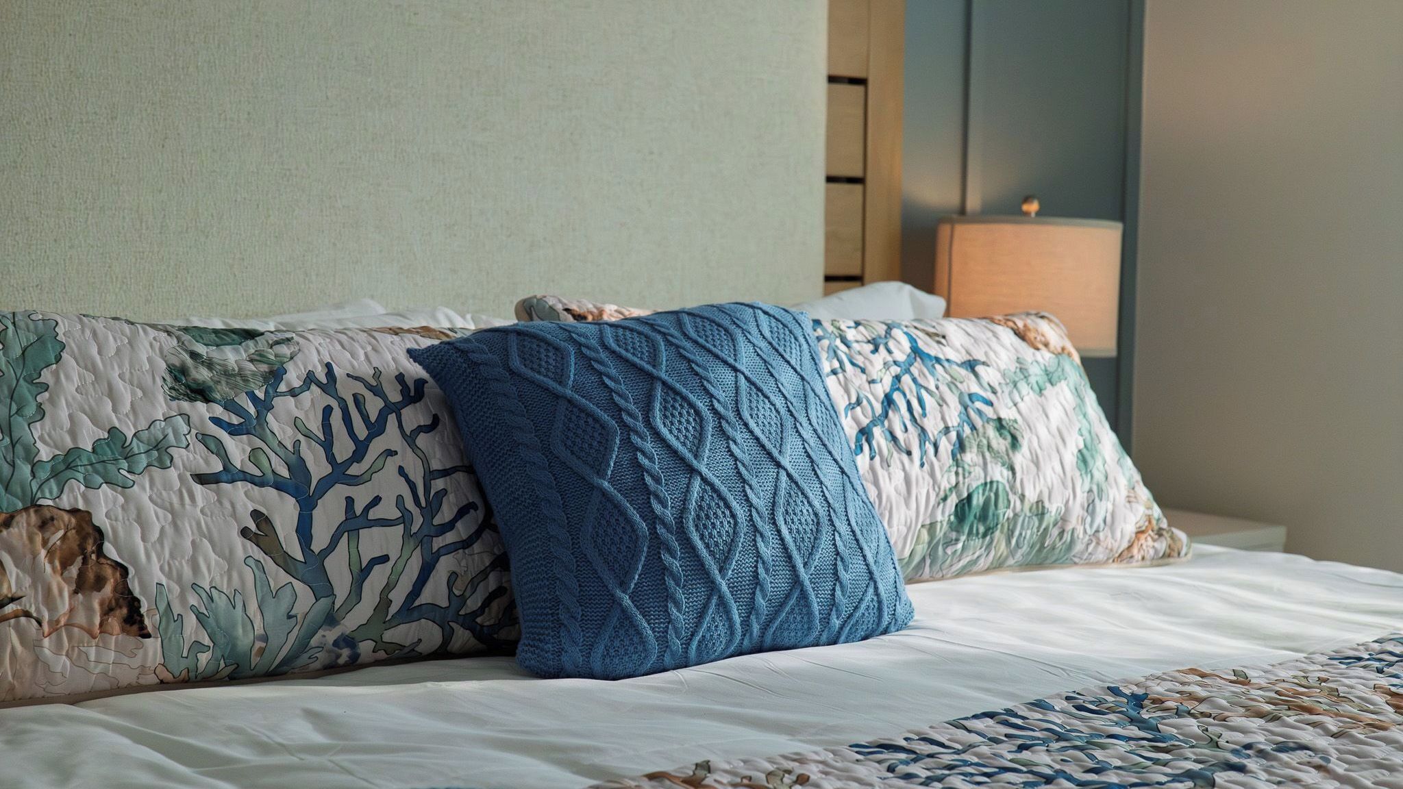A bed with a white sheet, two decorative pillows with a coral pattern, and a blue knitted pillow.