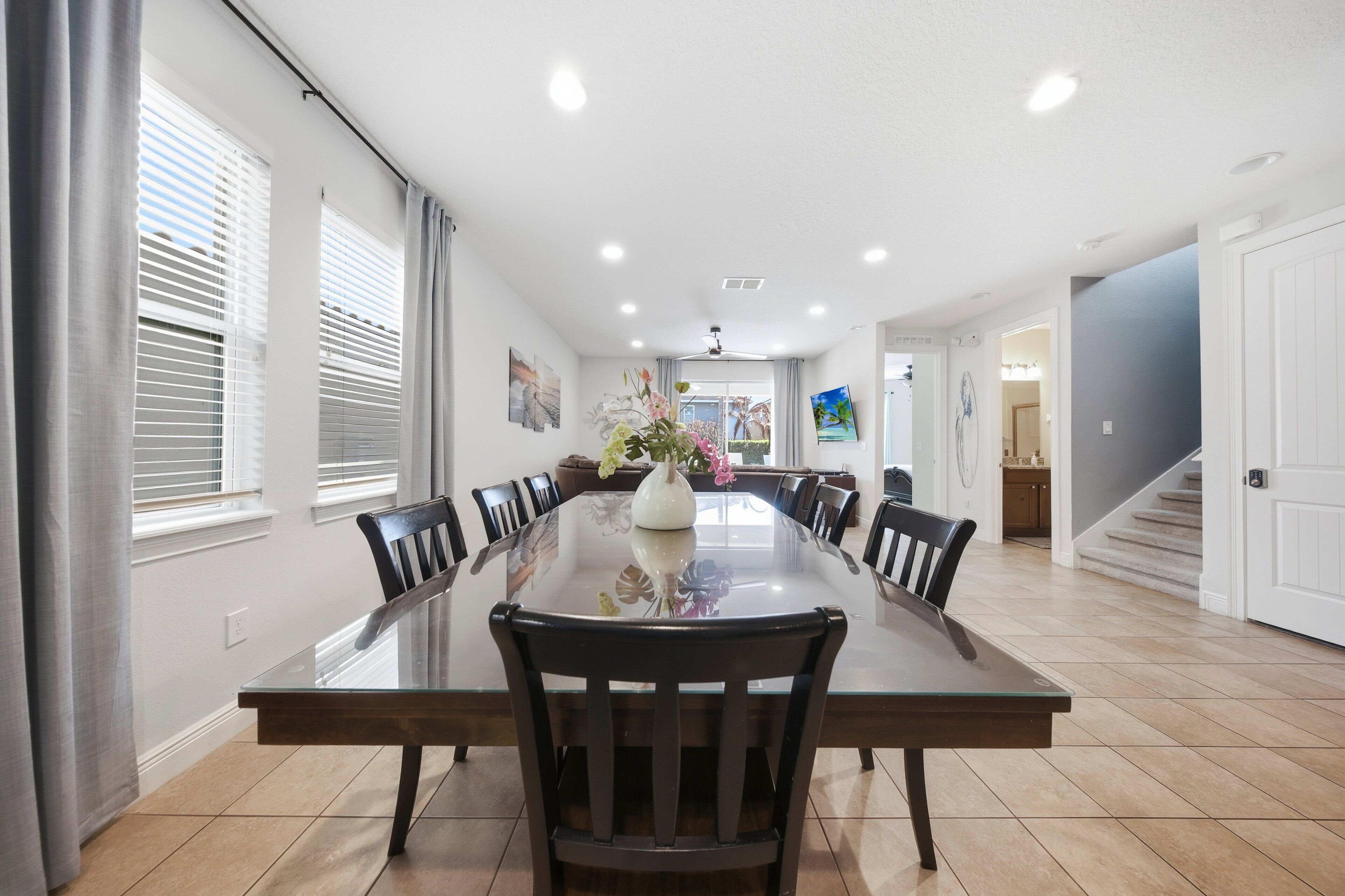 Une salle à manger avec une longue table, des chaises et un vase de fleurs. On y voit des fen êtres avec des stores, un ventilateur de plafond et un escalier au fond.