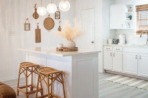 Uma cozinha com armários brancos, uma ilha central com banquetas, luminárias pendentes e adornos decorativos nas paredes.