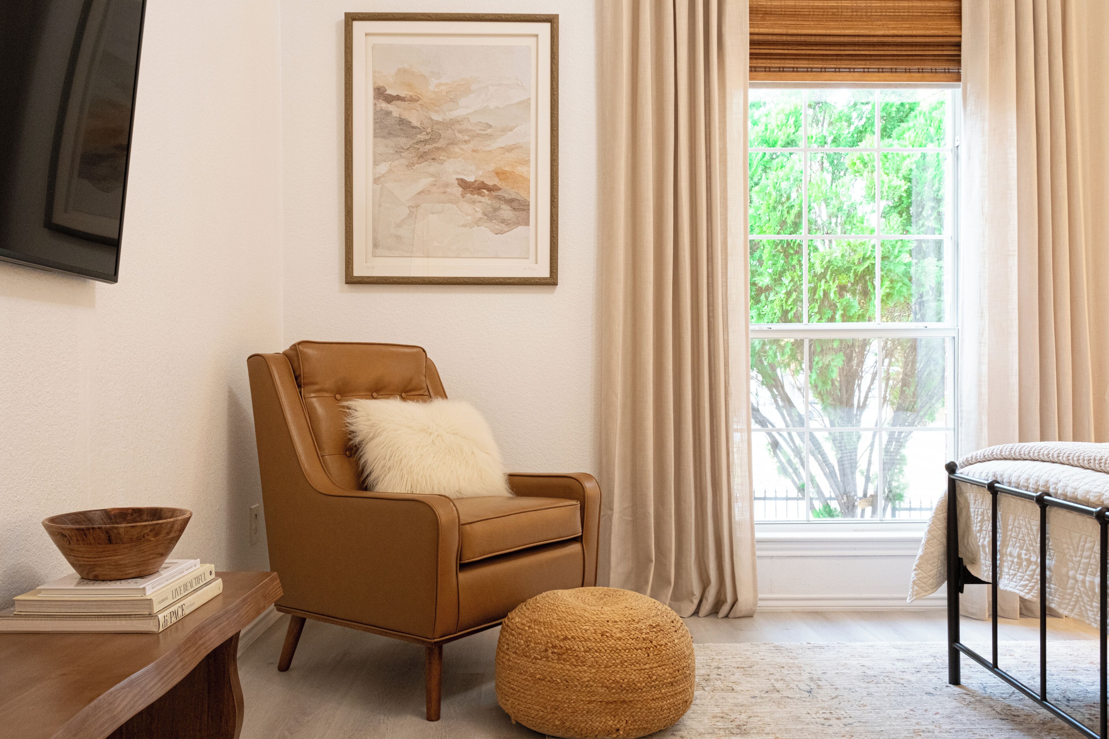 A room with a brown leather armchair, a round woven ottoman, a framed abstract painting, and a window with curtains.