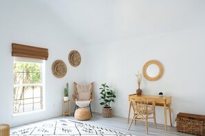 Een kamer met een groot raam, een houten bureau, een stoel, een klein tafeltje, een kamerplant en een gevlochten mand.