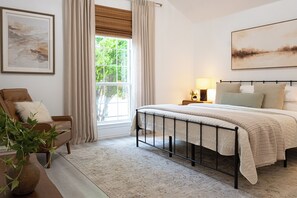 A bedroom with a bed, a nightstand, a lamp, a chair, a window with curtains, and a painting on the wall.