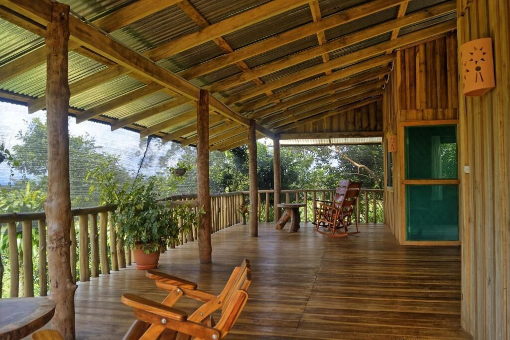 Una terraza de madera con bancos y una mesa, rodeada de plantas en macetas y una barandilla de madera.