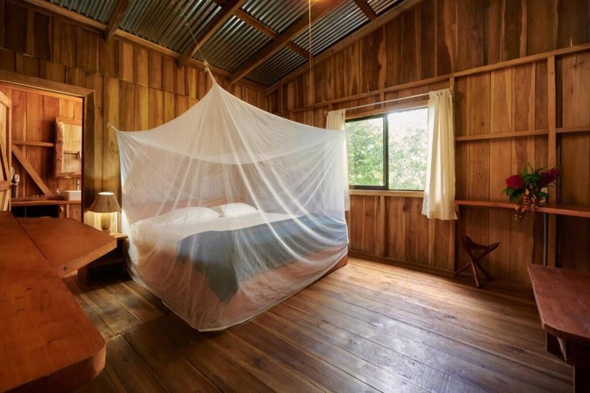 Een houten kamer met een bed onder een klamboe, een raam met gordijnen, en een houten bureau.