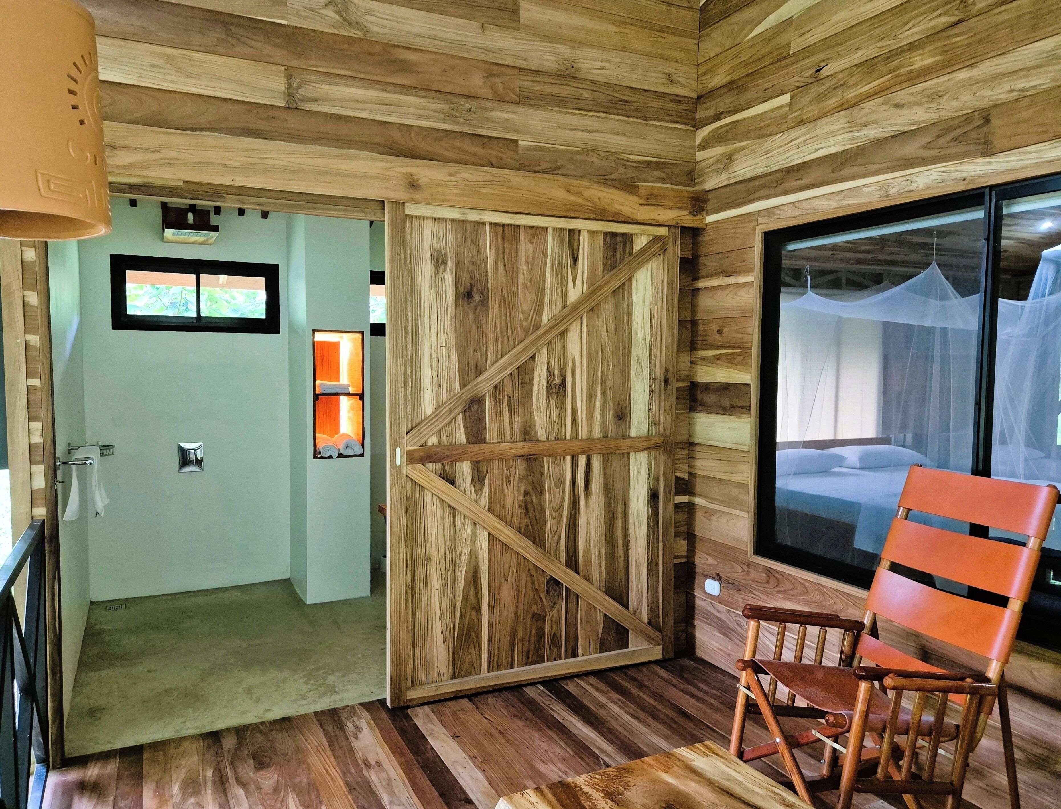 Un interior de madera con una puerta corredera que da a un baño, una cama con mosquitero y una silla de madera.