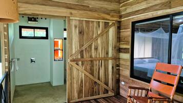 Un interior de madera con una puerta corredera que da a un baño, una cama con mosquitero y una silla de madera.