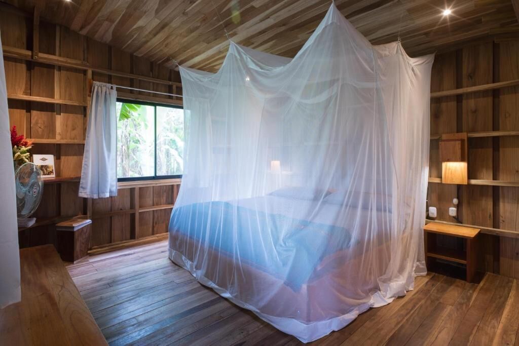 Ein Zimmer mit Holzboden und -wänden, einem Bett mit weißem Baldachin, einem Fenster mit Blick ins Grüne und einem Nachttisch mit Lampe.