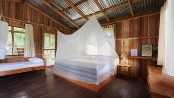Una cabaña de madera con una cama bajo una mosquitera, una mesita con una lámpara y una ventana con cortinas.