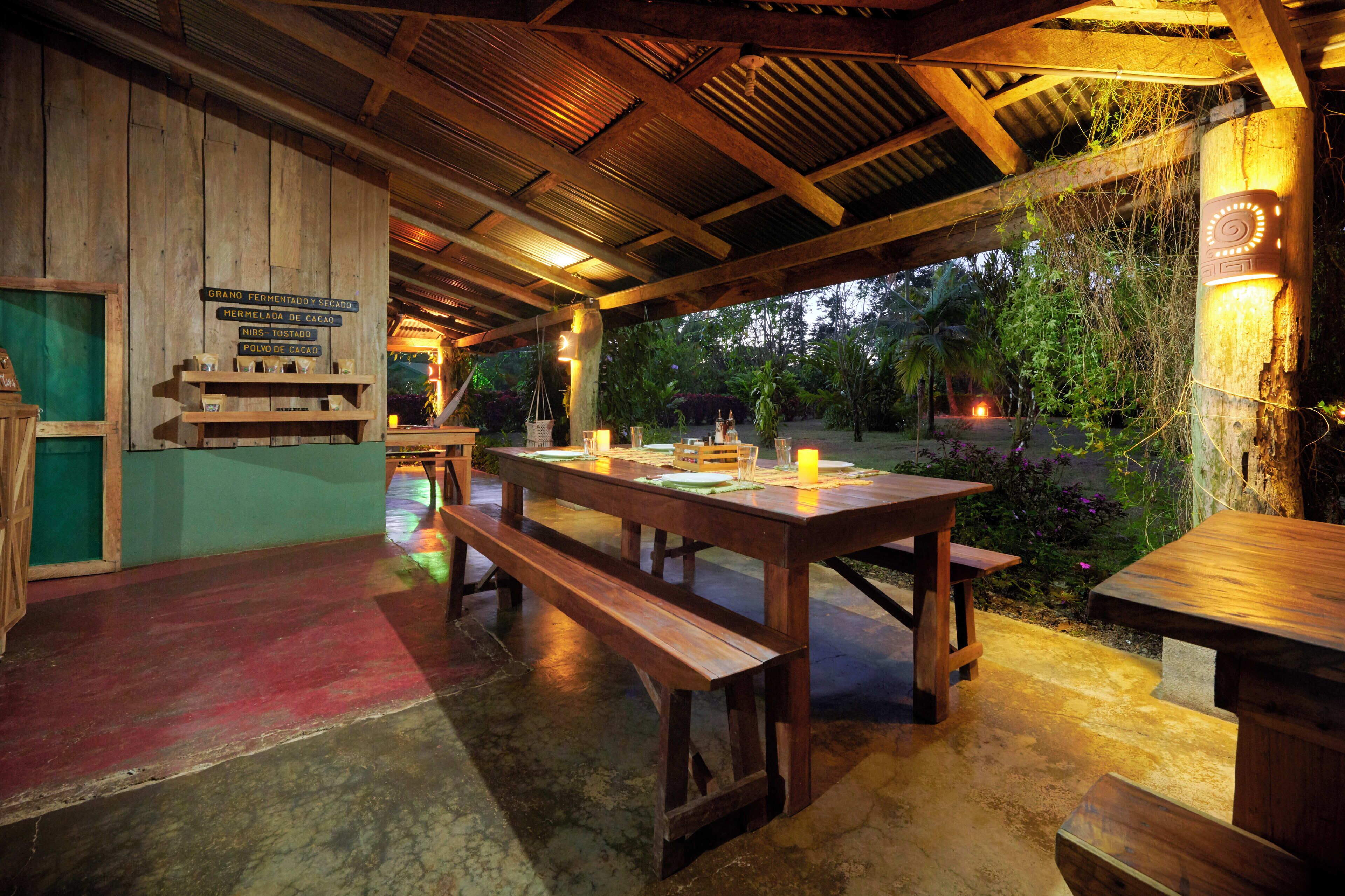 Un área de comedor al aire libre cubierta con bancos y mesas de madera, rodeada de follaje y un muro de madera con señalizaciones.