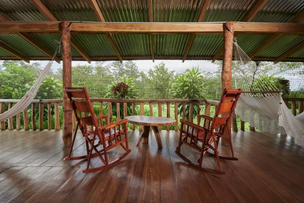 Una terraza de madera con una mesa y dos mecedoras, rodeada por una barandilla de madera y una hamaca.