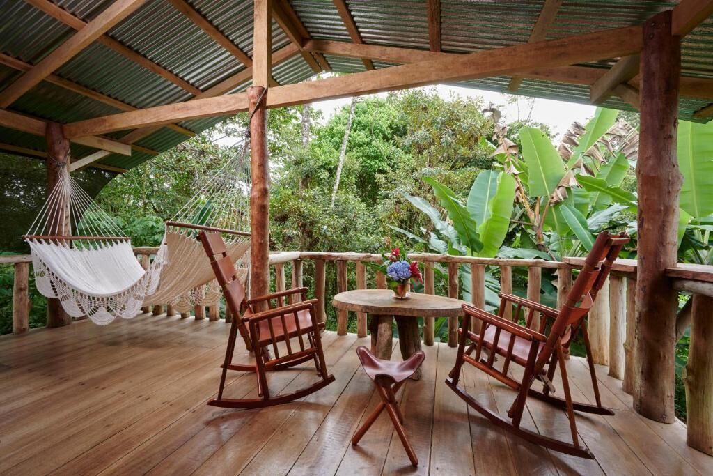 A wooden deck with a covered area, two rocking chairs, a small round table with a vase of flowers, and a hammock.