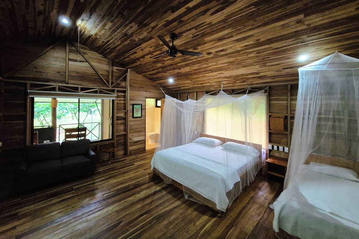 Een slaapkamer in een houten hut met een bed, een stoel, een plafondventilator en een raam met uitzicht op groen.
