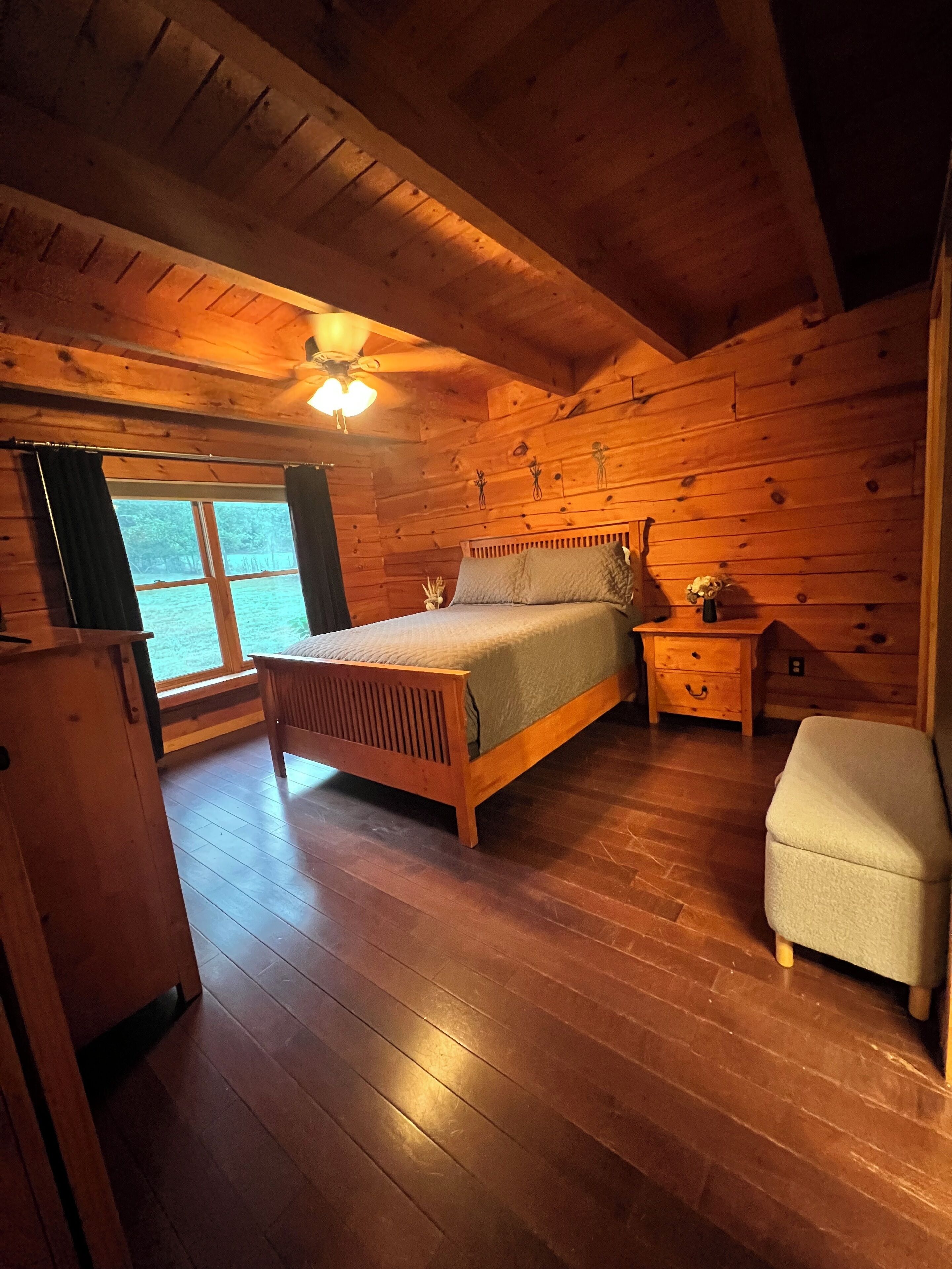A wooden cabin bedroom with a bed, bedside table, chair, and a window with curtains.