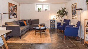 Una sala de estar con un sofá gris, sillones azules, una mesa de centro y una alfombra. Hay dos ventanas con plantas en el alféizar, una lámpara y una estantería llena de libros.