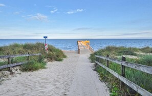 Ein Sandweg am Strand, der zu einem hölzernen Steg mit einer Rettungsboje und einem Schild führt.